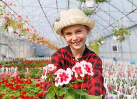 Junges Mädchen steht in einem Gewächshaus mit Blumen und hält eine Blume in die Kamera